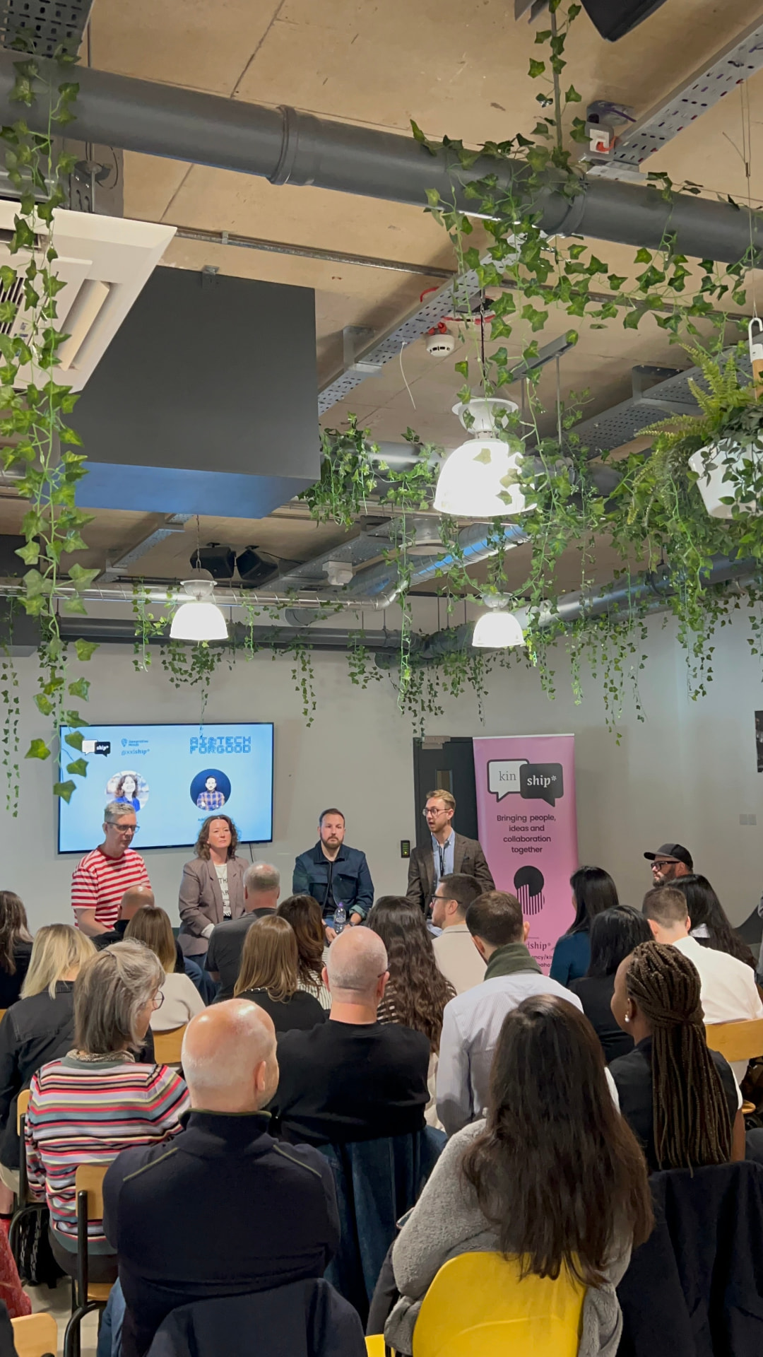 Panel discussion at a KinShip knowledge-sharing event, with speakers and attendees exchanging ideas beneath hanging plants